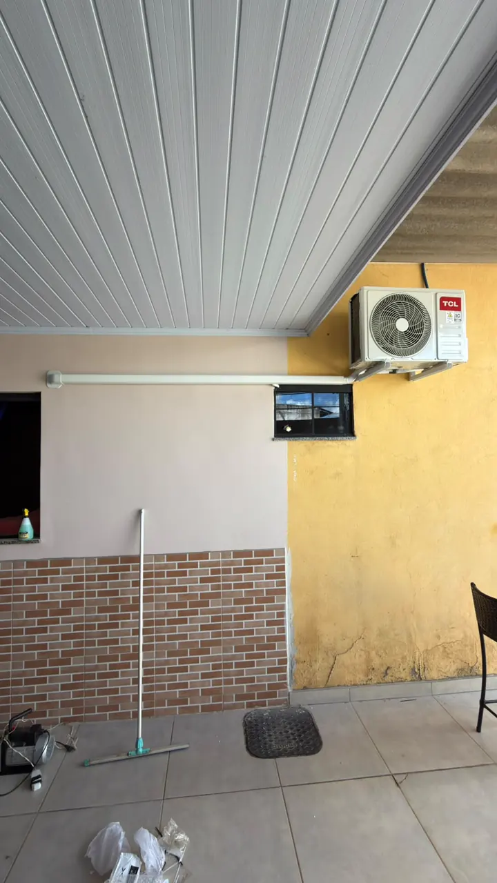 Condensadora TCL instalada no alto de uma parede amarela, numa área coberta com forro de PVC.
