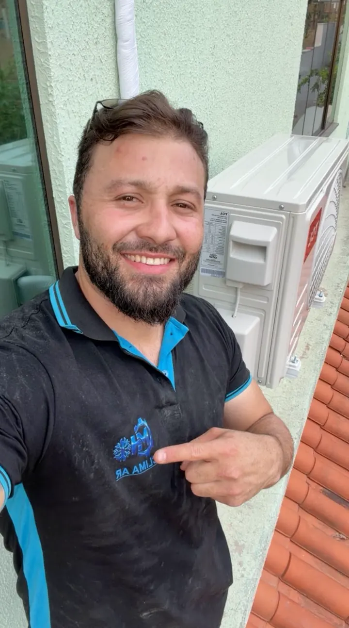 Mateus Henrique sorrindo, de camisa da MH Clima Ar, apontando para a marca bordada, ao lado de uma condensadora instalada.